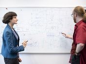 Eine Forscherin (links) und ein Forscher (rechts) stehen vor einem dicht beschriebenen Whiteboard und diskutieren eine Forschungsfrage.