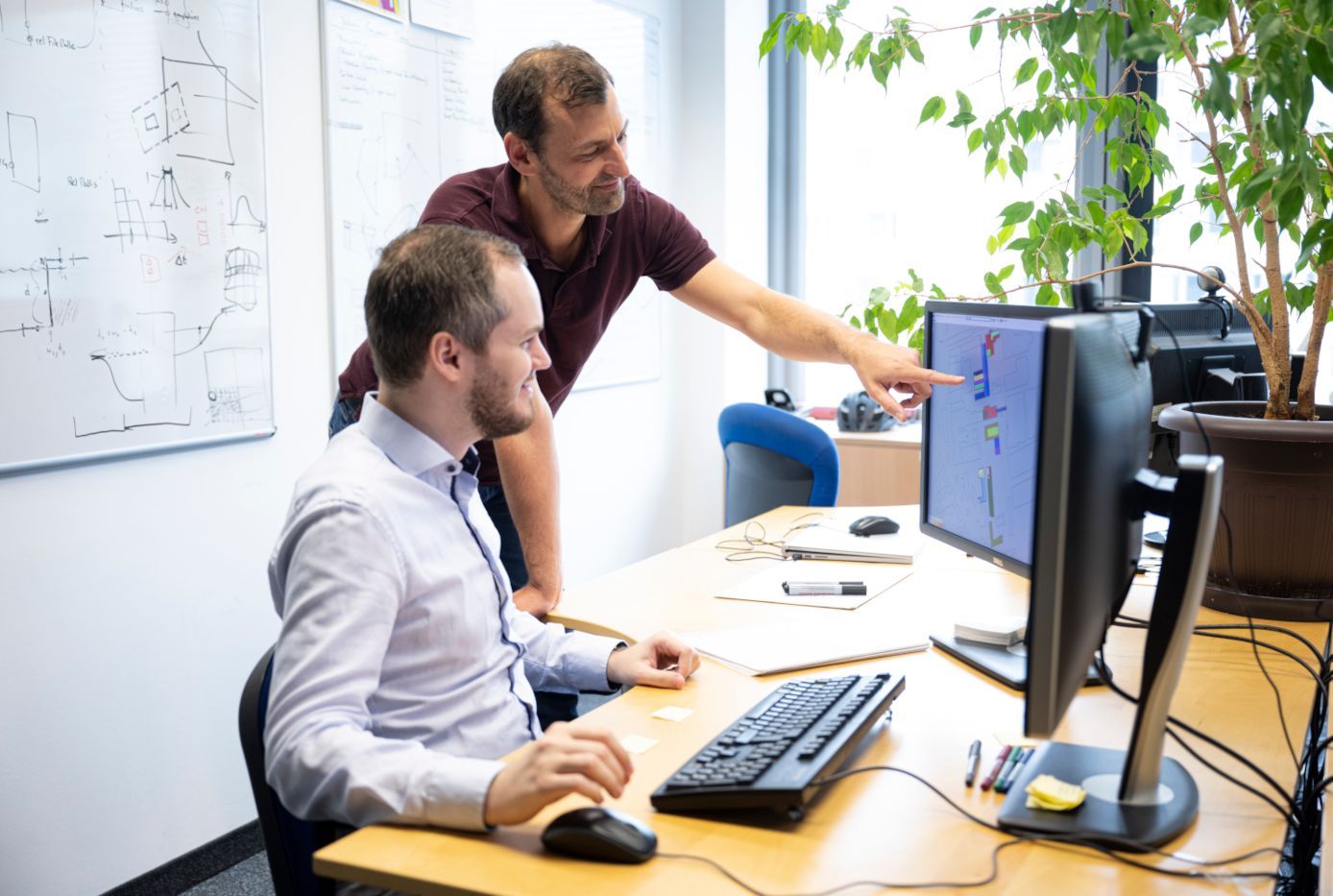 Zwei Forscher des VRVis arbeiten gemeinsam an einem Desktop-Computer. Ein Forscher sitzt, der andere steht und deutet auf den Bildschirm. Im Hintergrund ist ein Whiteboard mit Forschungsnotizen zu sehen.
