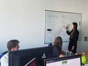 Two students have a researcher explain something to them on the whiteboard. 